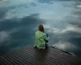 Eine Frau sitzt allein auf einem Steg, blickt aufs Wasser. Die Spiegelung der Wolken im Wasser verstärkt das Gefühl der **Angst vor dem Alleinsein**.