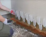 Osoba w rękawiczkach ustawia rurki przy ścianie, pokazując, jak osuszyć ściany domowym sposobem.