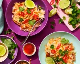 Två skålar med kyckling pad thai, toppad med chili, koriander och jordnötter. Limeklyftor och en liten skål med chilisås finns bredvid.