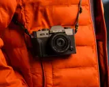 Srebrno-czarny aparat Fujifilm X-T30 z obiektywem, zawieszony na pasku na tle pomarańczowej, pikowanej kurtki. Idealny aparat na wakacje, gotowy do łapania wspomnień.