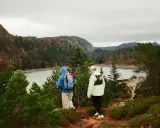 Två personer vandrar längs en stig med en sjö och berg i bakgrunden. Perfekt för att upptäcka Sveriges vandringsleder.
