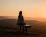 Eine Person sitzt auf einer Bank und blickt in die Ferne. Die untergehende Sonne taucht die Hügellandschaft in warmes Licht, ein Moment, um im Reinen zu sein.