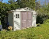 Un abri de jardin en plastique beige, avec des fenêtres et une double porte, est prêt pour son aménagement. Un petit panneau solaire est installé sur le toit.