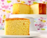 Un delicioso bizcocho de yogur, esponjoso y dorado, servido en un plato blanco. Otro trozo se ve en un soporte para pasteles al fondo.