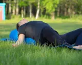 Person ligger på en blå yogarulle i gräset, med böjda knän och armarna utåt. En lugn stund med yin yoga utomhus.