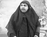 Zahra Khanom Tadj es-Saltaneh, księżniczka perska, w czarnym hidżabie i pasiastym stroju, pozuje na tle ogrodu.
