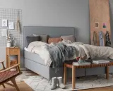 Ett mysigt sovrum grått med en stor säng, en trästol och en sittbänk. En rutig väggdekoration med bilder och en vas med bomull.