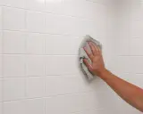 Mano limpiando azulejos blancos recién colocados con un paño gris.
