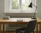 Drewniane biurko pod oknem z kaloryferem. Na blacie notatnik, długopis i lampka. Obok krzesło.