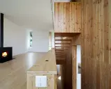 Nowoczesne wnętrze z kominkiem i schodami. Dominują jasne drewno i boazeria ze sklejki, tworząc ciepłą atmosferę.