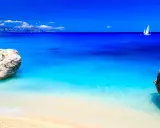 Spiaggia con rocce e mare cristallino, un veliero solca le acque turchesi. Potrebbe essere il mare più bello in Italia.