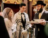 Novios sonríen durante su **matrimonio judío**. El novio lleva un talit y el rabino lee un documento.