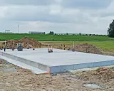 Gotowe fundamenty krok po kroku. Płyta betonowa na budowie, otoczona piaskiem i zielenią. W tle widać budynki przemysłowe.