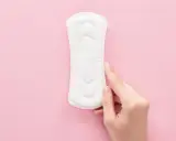Una mano toca una compresa sobre fondo rosa. Representa cómo es el flujo antes de la regla, ligero y discreto.