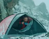 Mężczyzna w namiocie, jedzący posiłek, w zimowej scenerii górskiej. Scena jak z filmów wspinaczkowych.