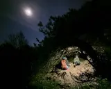 Två personer sitter under en grenig gran, redo att sova under bar himmel. Månen lyser upp natthimlen.