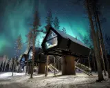 Hus på styltor i snöig skog under norrsken. En **arctic road** leder fram till stugorna, upplysta av stjärnor och lampor.