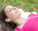 Kobieta leży na trawie, słuchając muzyki. Widać jej twarz z zamkniętymi oczami i uśmiechem. To dowód na to, jak wielki wpływ muzyki na psychikę człowieka.