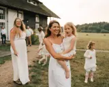 Rodzina w białych strojach, wśród nich kobieta z córką na rękach. Tło stanowi stodoła i pole. To był skromny ślub kościelny bez wesela.