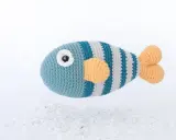 Un pez amigurumi a rayas azules y grises con aletas naranjas, flotando sobre guijarros blancos. ¡Encuentra aquí peces amigurumi patrón gratis!
