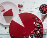Ein köstlicher no bake Himbeer Cheesecake, dekoriert mit frischen Beeren und Schokoharz. Ein Stück ist bereits auf einem Teller.