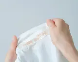Dłonie trzymają białą bluzę z plamami podkładu. Dowiedz się, jak zmyć podkład z bluzy.