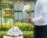 Mężczyzna w białej koszuli i niebieskim krawacie trzyma bukiet białych kwiatów i różaniec. To jego urlop okolicznościowy śmierć bliskiej osoby.
