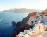 Traumhafter Blick auf die weißen Häuser und blauen Kuppeln von Santorini. Das Meer glitzert, Boote fahren vorbei. Ein perfekter Santorini Strand Urlaub.