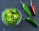 Frisch geschnittene Jalapeños in einem Glas, bereit zum Jalapenos einlegen. Daneben liegen drei ganze Chilis, eine davon reif rot.