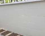 Mur extérieur en béton cellulaire peint en blanc, avec une bordure en pierre grise au sommet. Des plantes poussent à la base.