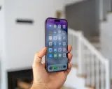 Dłoń trzyma smartfon z widocznym ekranem iPhone 14 Pro Max. Na wyświetlaczu widać ikony aplikacji i godzinę 11:02.