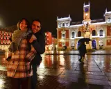 Pareja sonriente en la Plaza Mayor de Valladolid, disfrutando de las atracciones nocturnas y el reflejo de las luces en el suelo mojado.