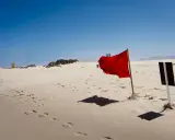 Pusta plaża z piaskiem, na którym widać ślady stóp. W oddali żółta budka ratownicza, a na pierwszym planie powiewają czerwone flagi ostrzegawcze.