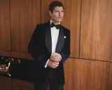 A man in a sophisticated navy velvet tuxedo, embodying perfect black tie attire, stands by a modern fireplace.