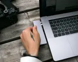 Dłoń z tatuażem pioruna podłącza adapter do laptopa, obok leży aparat. Idealny zestaw, jaki laptop do obróbki zdjęć?