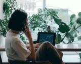 Kobieta w białej koszulce rozmawia przez telefon, pracując na laptopie z wykresem giełdowym. To jej wolny zawód, który pozwala jej pracować w otoczeniu zieleni.