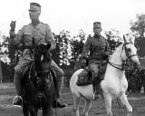 Två militärer till häst, kanske under en övning för regementen i Sverige. Bakom syns fler soldater och träd.