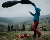 Osoba w niebieskiej kurtce i czerwonych spodniach rozkłada śpiwór puchowy czy syntetyczny na tle górskiego krajobrazu.