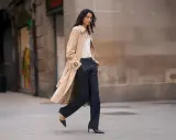 A woman walks confidently in dark palazzo pants and a trench coat, proving they are still in style.