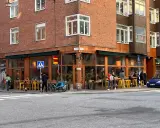 Ett mysigt hörnkafé i **Södermalm, Stockholm** med folk som sitter vid gula stolar och bord utomhus. En blå cykel med barnvagn står parkerad.
