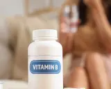 Biała butelka z etykietą "VITAMIN B" na pierwszym planie. W tle niewyraźna postać kobiety z kieliszkiem wody, sugerująca potencjalny efekt niedoboru witaminy B1.