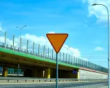 Pomarańczowy znak "ustąp pierwszeństwa" ostrzega przed wjazdem na drogę podporządkowaną. Pod wiaduktem widać drogę i słupy oświetleniowe.