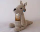 Canguro amigurumi beige sentado, con ganchillos en su bolsa y marcadores de puntos en su oreja.