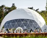 Una gran cúpula estructura blanca, con ventanas triangulares, se alza en un día soleado. Gente pasea frente a ella, rodeada de árboles verdes.