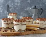 Plusieurs verrines de tiramisu sans café, garnies de cacao, côtoient des biscuits à la cuillère et des grains de café sur une planche en bois.