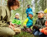 En grupp barn och en vuxen njuter av en dag i mulle skogen, omgivna av träd och grönskande natur.