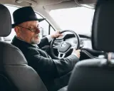 Starszy pan w kapeluszu i okularach prowadzi samochód. W ręku trzyma dowód rejestracyjny, który jest dowodem współwłaściciel.