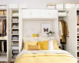 Habitación matrimonio pequeña con armario blanco, cama con ropa de cama amarilla y almohadas, y estanterías llenas de ropa y cestas.