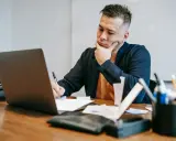 Mężczyzna zamyślony nad notatkami przy laptopie. Jego progresywny styl i skupienie sugerują pracę nad ważnym projektem.