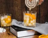 Ein Old Fashioned wird zubereitet, Eiswürfel und Orangenschale zieren das Glas. Ein altes Rezept wird lebendig.
